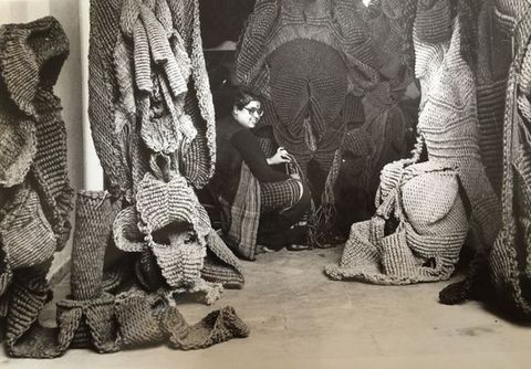 Black and white photograph. A woman sits in a studio draped with woven sculptures.