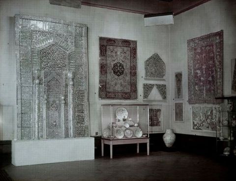 Colour transparency. Textiles hang on walls around a case filled with dishes on three stepped levels. A two-storey silver architectural feature is elaborately patterned.