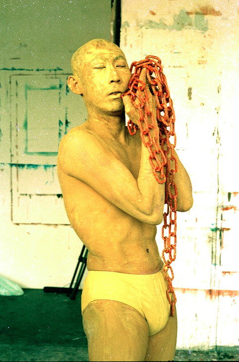 Performance still. The skin of a bald man wearing only yellow briefs is painted yellow. He holds up a handful of thick, red chains by his face, his eyes closed.