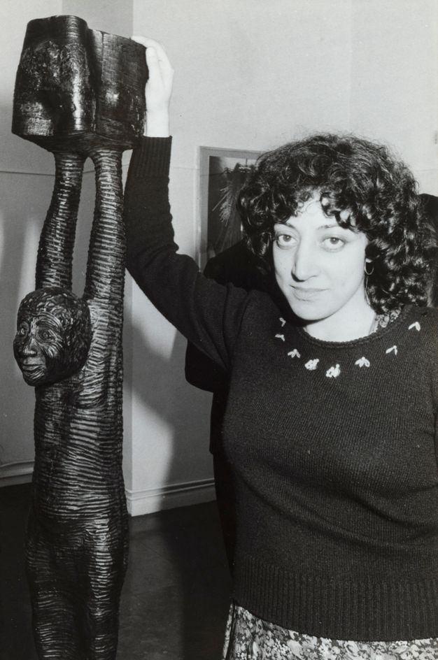 Black and white photograph. A woman with black curly hair falling her shoulders stands next a sculpture as she looks at us. The sculpture is of a person with their arms raised. She rests one hand on the top, which comes up over her head.
