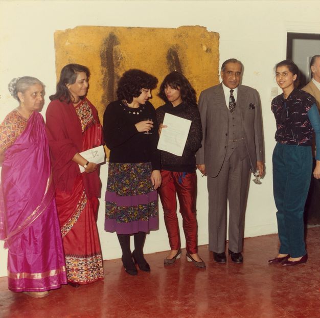 Five women and a man with medium skin stand in a row in a gallery hung with works of art.