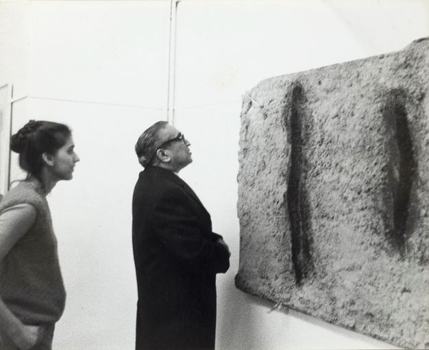 Installation. Black and white photograph of a man and woman looking at a sculptural piece hanging on a gallery wall.