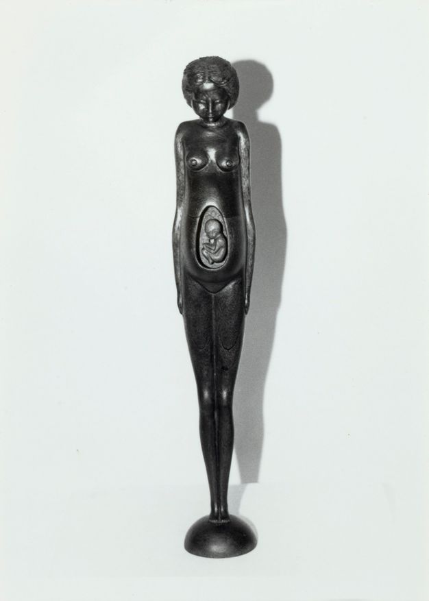 Sculpture. Black and white photograph of a nude woman stands with her arms by her side and small feet together, creating a sliver-like silhouette. An oval section of her torso has been removed to show a foetus in her womb.