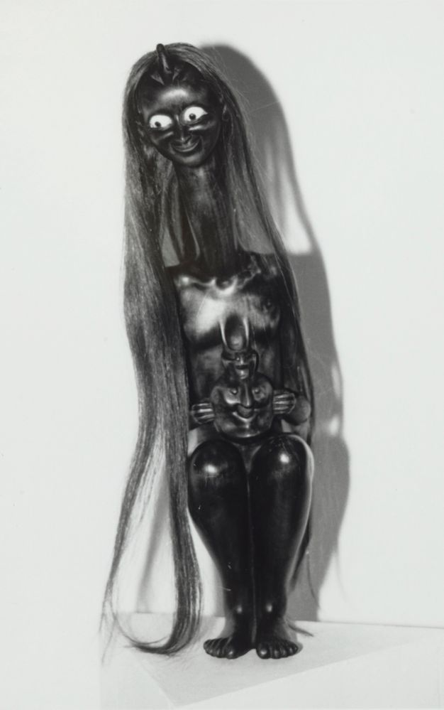 Sculpture. Black and white photograph of a sitting woman with a serpentine neck. Her head angles forwards and to our left, long hair affixed to the crown falling to her feet. Her bulging eyes are white with black pupils. The rest of her body is polished black.