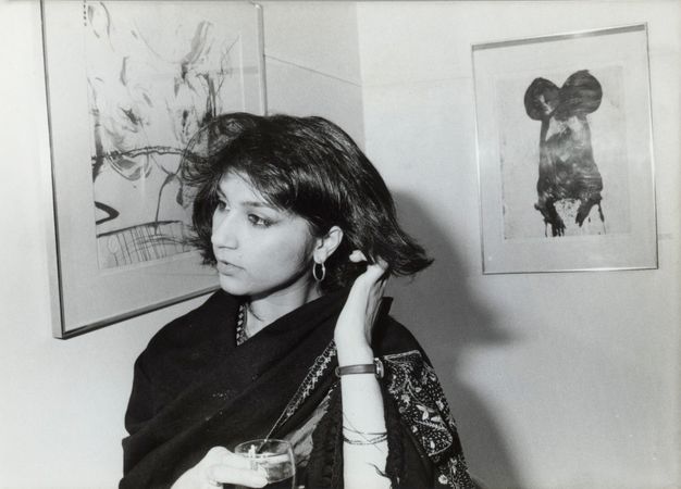 Black and white photograph. A woman with chin-length black hair stands in front of two prints hanging in a corner. A scarf or garment wraps around her shoulders, and she holds a glass in one hand.