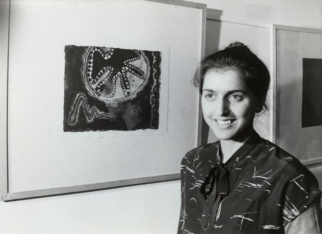 Black and white photograph. A woman with her black hair pulled back stands smiling next to an abstract framed painting. A black shirt with light-coloured slashes ties in a bow at her neck.