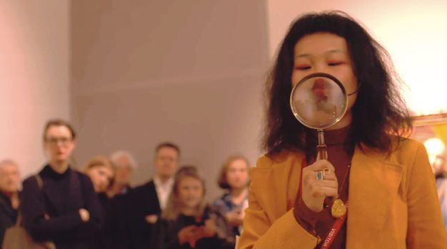 Performance still. A medium-skinned woman with black, shoulder-length hair, purses her lips behind a magnifying glass she holds up to her face. Onlookers are blurry in the background.