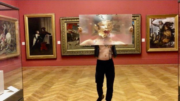 Performance still. A medium-skinned holds a sheet of magnifying plastic, arms stretched wide, as he walks towards us a gallery hung with paintings in gold frames. His face is covered in gold leaf, and his mouth is filled with red chili peppers.