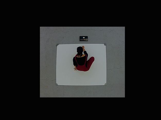Video. From above, we watch a person with dark hair draw a circle, spinning on one foot to enclose their body, onto a piece of paper affixed to a grey concrete floor. The video plays on a loop.