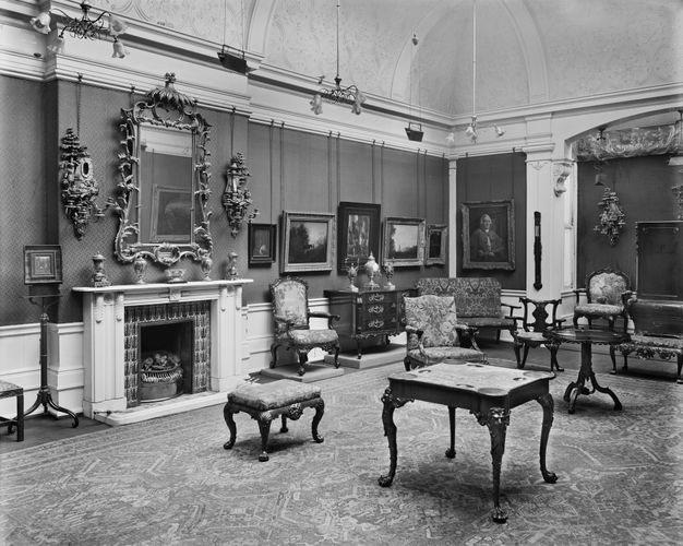 Black and white photograph. A room is hung with paintings and decorated with vases, a gilded frame, and antique furniture. Panelling is below a chair rail, and thick moulding lines the door. The ceiling curves up over the crown moulding and is decorated with plaster floral designs.