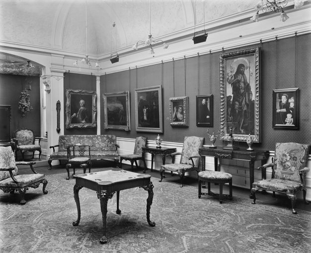 Black and white photograph. A room is hung with paintings and decorated with vases, statuettes, and antique furniture. Panelling is below a chair rail, and thick moulding lines the door. The ceiling curves up over the crown moulding and is decorated with plaster floral designs.