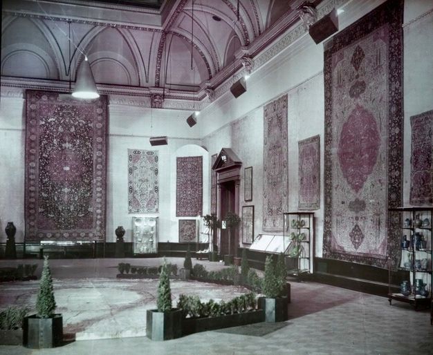 Colour transparency. Rugs are hung closely together on stone walls under elaborate moulding. The ceiling has lunettes set into curving arches lined with gilded carving. Two rugs reach nearly from the floor to the moulding around the high ceiling.
