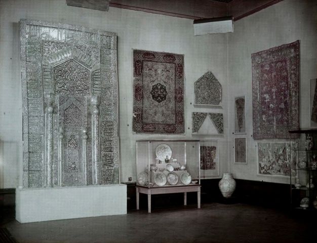 Colour transparency. Textiles hang on walls around a case filled with dishes on three stepped levels. A two-storey silver architectural feature is elaborately patterned.