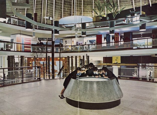 Installation. Seven people gather around the round surface of a light table at the centre of a three-storey atrium.