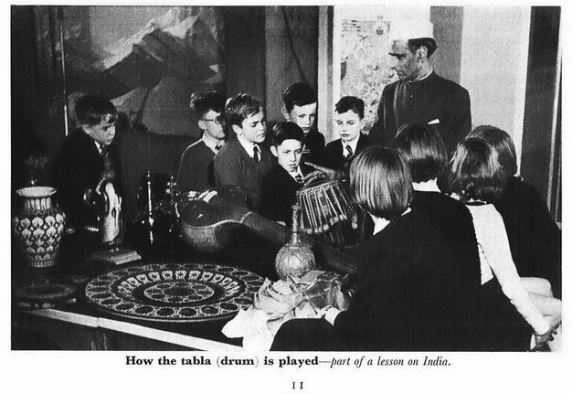 Illustration. Black and white illustration shows a group of children wearing suits or dresses with a drum. A man wearing a white cap and high-necked dark garment stands at the back of the group. Text below reads, “How the tabla (drum) is played – part of a lesson on India”.