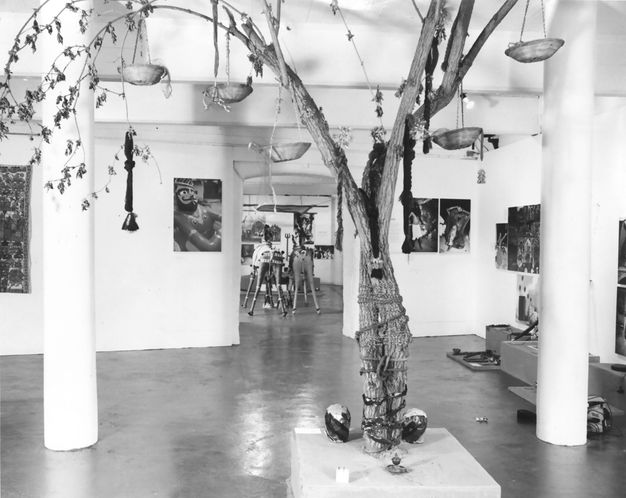 Installation. Black and white photograph of bowls hanging from a tree in a gallery space. The trunk is bound, or it might tie two trunks together. Works of art are on the walls and more sculpture is through a doorway opposite us.