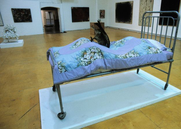 Installation. A metal bedstead on wheels is on a low white platform in a gallery hung with works of art. A thin mattress on the bed is covered with a blue, floral blanket, which bulges as if over a body. A large black bird dives into the bed, headfirst into where the gut or groin of the person lying there would be.