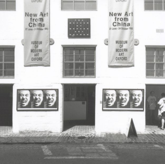 Black and white photograph. Two banners hanging on a white building have the title and location of the exhibition along with the dates, “27 June – 20 October 1993”. Below each hangs a panel showing a close up of a laughing face three times. People enter a doorway to our right.