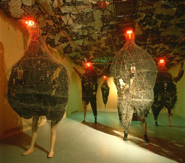 Installation. Five sculptural forms stand in a long gallery space. Each has the legs of a mannequin and bodies that are either humanoid or shaped more like a chicken. Each body has black-coloured material encased in chicken wire, and red lights glow like eyes from the very tops of tiny heads.