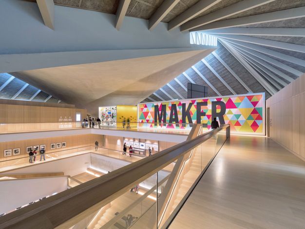 Architecture. At least four storeys open onto a central space under an angular ceiling. We are on the top floor, across from an exhibition sign reading “Maker”. The floors on all levels are blond wood, and some of the wood walls are lined with works of art.