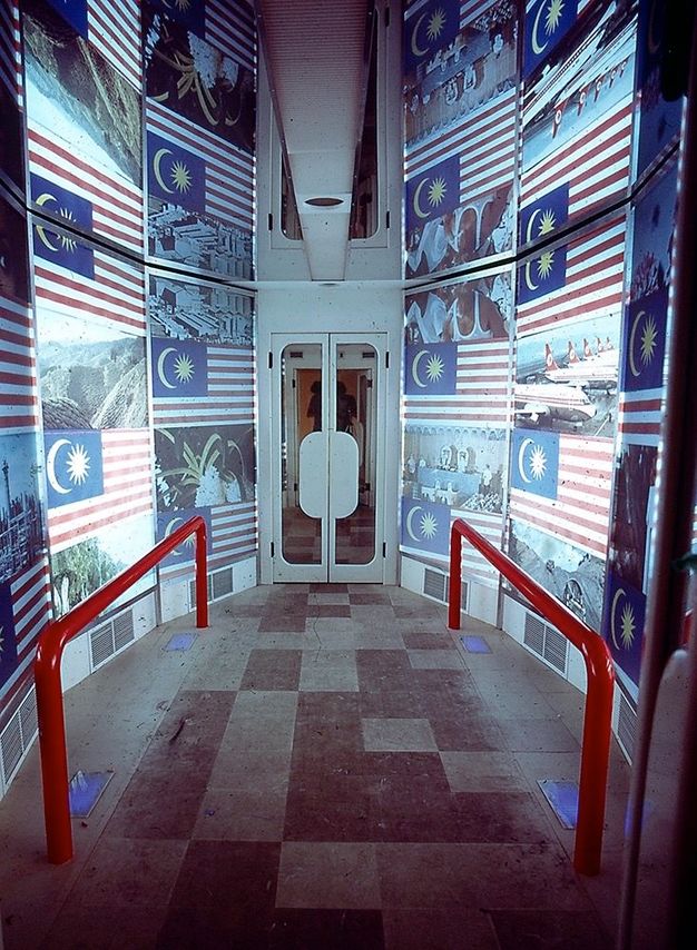 Installation. A walkway flanked by red railings lead to double doors. The walls are covered with the Malaysian flag alternating with photographs.