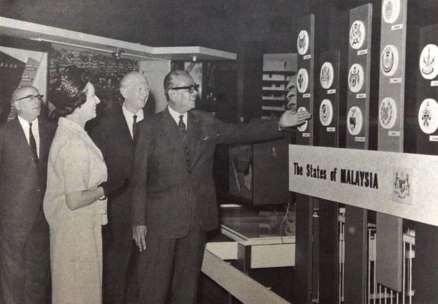 Black and white photograph. A man wearing glasses and smiling holds out a hand to gesture towards a display illustrating “The States of Malaysia”. Two other men and a woman look on, two of them also smiling. The men wear suits, and the woman a pillbox hat and gloves, with a skirt suit.
