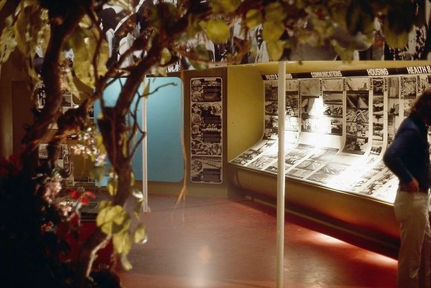 Installation. Inside a gallery, we look through tree branches at a display of black and white photographs that covers the far wall and curves down and towards us, making a low, angled counter. The headings over the images that we can make out read “Communications”, “Housing”, “Health &”, with the final one cut off.
