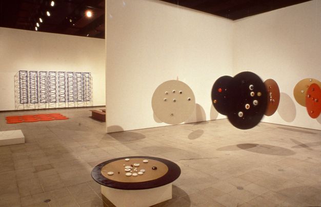 Installation. Six round discs, like tabletops, float suspended from a gallery ceiling. A seventh sits on a pedestal and holds round stones or objects made of another material. The hanging discs are painted brown, red, or black.