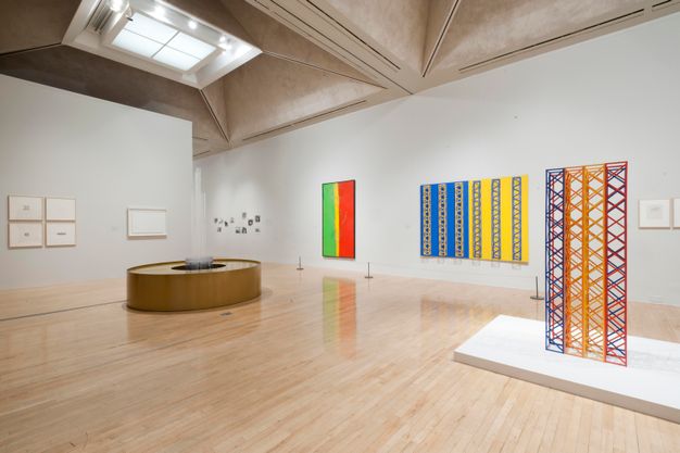 Installation. Two sculptures are in the centre of a gallery hung with mostly colourful works of art, many of which create patterns. The gallery itself has shiny wood floors and trapezoid-shaped coffers set into the ceiling.