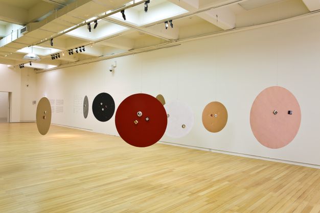 Installation. Eight discs like tabletops are suspended from the ceiling. The discs are blood red, rose pink, white, black, or tan. Two or three round or square objects are affixed to the faces of the discs.