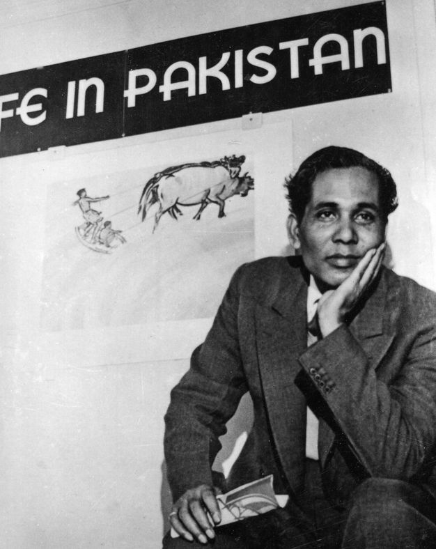 Black and white photograph. A man sits with his head resting in one hand, that elbow propped on a knee as he looks off to our right. He has dark skin and black hair, dark brows, and rounded features. A drawing of two animals pulling two people on a sled hangs on the wall behind him, under the exhibition sign.