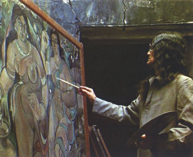 Video still. A medium-skinned man with long black hair painting a canvas showing two life-sized, stylized, medium-skinned women. He holds a round palette in the hand closer to us.