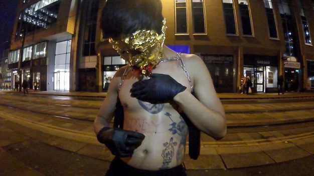 Performance still. On a street at night, a medium-skinned, bare-chested man cuts letters into the skin of his abdomen. He wears black gloves, and his face is covered in gold leaf.
