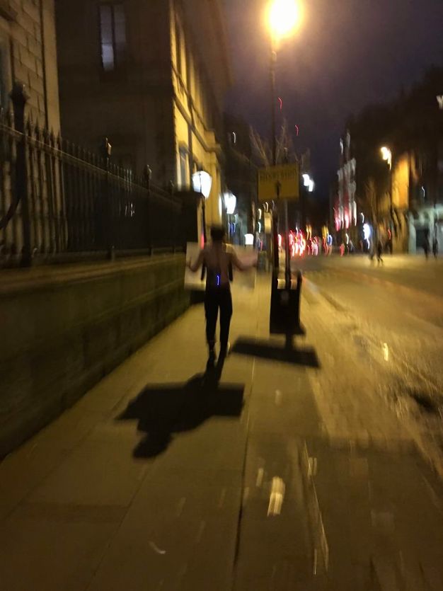 Performance still. A bare-chested man walks away from us, holding an object in front of him. The photograph is very blurry.