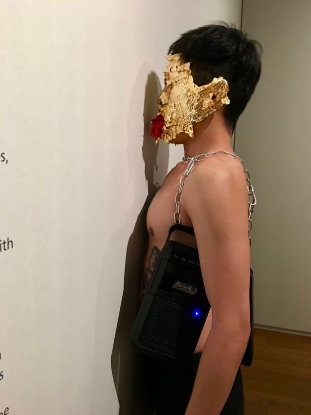 Performance still. A medium-skinned, bare-chested man presses his torso to a gallery wall. Cassette players hang from chains around his chest, and his face is covered with gold leaf. Red chili peppers flick out of his mouth like thin tongues.