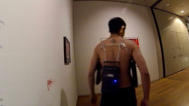 Performance still. A medium-skinned, bare-chested man walks away from a bloody mark on a gallery wall. Cassette players hang from chains around his torso.