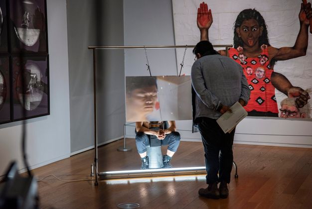 Installation. A person stands looking at a performance art piece. The artist sits on an upturned metal bucket with a magnifying sheet of plastic, the size of a large TV screen, which hangs from a metal frame down in front of his face. The artist’s eyes are closed and fingers interlaced, elbows resting on knees.