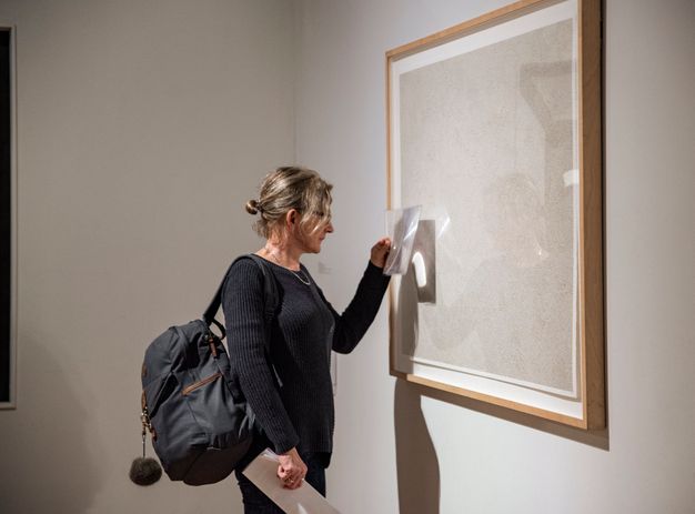 A woman holds a sheet of magnifying material, about the size of a tablet, up to a framed, white work of art, looking closely at the surface.