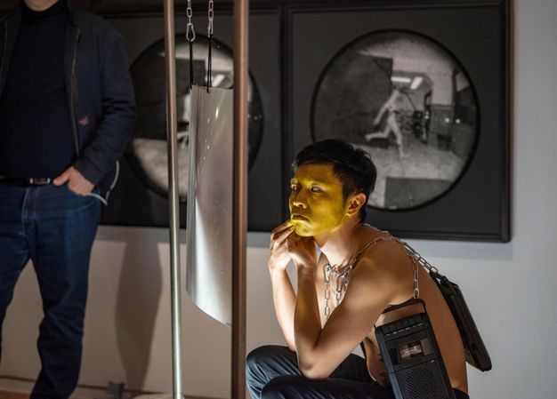 Performance still. A medium-skinned man applies yellow paint to his face. A sheet of magnifying plastic hangs from a metal frame to our left. Cassette tape players hang from chains around the artist’s bare torso.