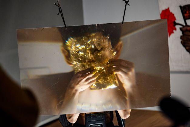 Performance still. In a gallery and from behind a sheet of magnifying plastic, the size of a large TV screen, an artist covers his face in gold leaf.