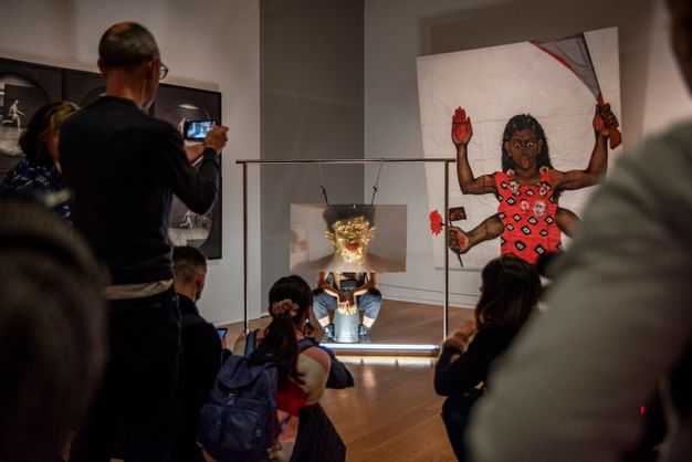 Performance still. A medium-skinned man sits on an upturned metal bucket in a gallery. He sits behind a sheet of magnifying plastic, the size of a large TV screen. He has covered his face in gold leaf and his mouth is full of whole red chili peppers, which stick out like tongues. People look on and record or take pictures, us included.
