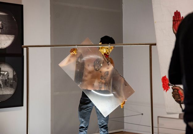 Performance still. A medium-skinned man removes a sheet of magnifying plastic, the size of a large TV screen, from a metal frame. The man’s face is covered in gold leaf and his mouth filled with red chili peppers.