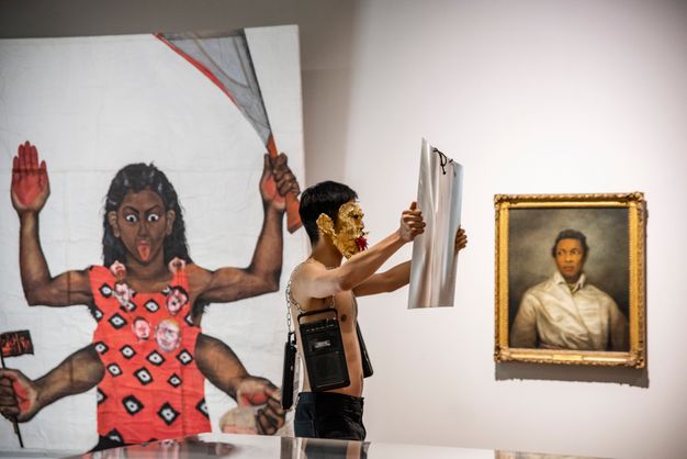 Performance still. A medium-skinned holds a sheet of magnifying plastic, arms stretched wide, as he walks through a museum. Cassette players hang from chains around his bare chest. His face is covered in gold leaf, and his mouth is filled with red chili peppers.