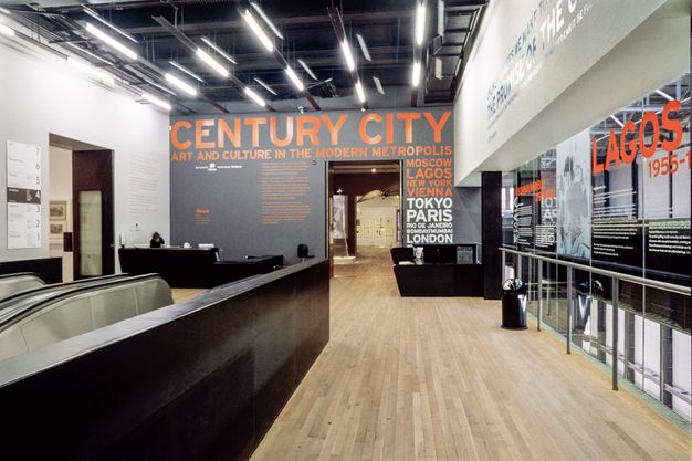 Installation. Two escalators lead up to the entrance of an exhibition. The title is in orange text on a black wall. Cities are listed down one side: Moscow, Lagos, New York, Vienna, Tokyo, Paris, Rio de Janeiro, Bombay/Mumbai, and London.