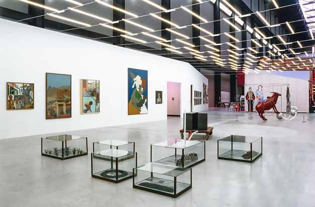 Installation. Six cases holding sculptures sit on the grey concrete floor of this gallery. Other sculptures without cases are in the space, and works of art line the walls.