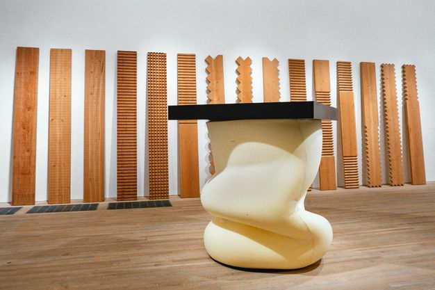 Installation. Fifteen wood planks leaning against the wall are carved with geometric patterns on the front faces or notched down the sides. A sculpture sitting on the floor has a black panel balanced on a folded form, like a slumped futon mattress or oozing bread dough.
