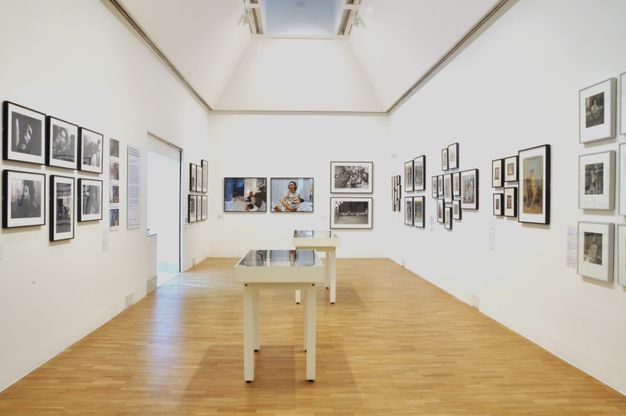 Installation. Framed black and white and colour photographs and perhaps other works on paper hang along the three walls of a gallery stretching before us. Two cases are in the middle of the space.