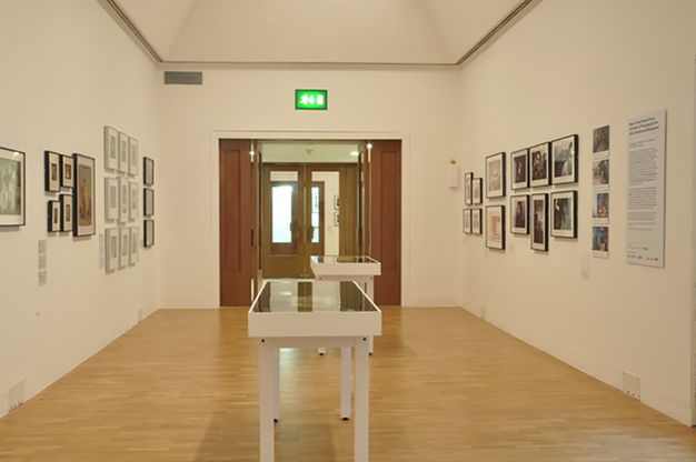 Installation. Framed black and white and colour photographs and perhaps other works on paper hang along two walls leading back to a doorway. Two cases are in the middle of the space.
