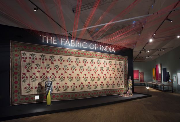 Installation. Letters spelling the name of the exhibition are made of white dots over a display of a long textile, perhaps ten paces across. Red threads extend from the dots of the title to points on the ceiling above us, so the threads make a starburst-like pattern.