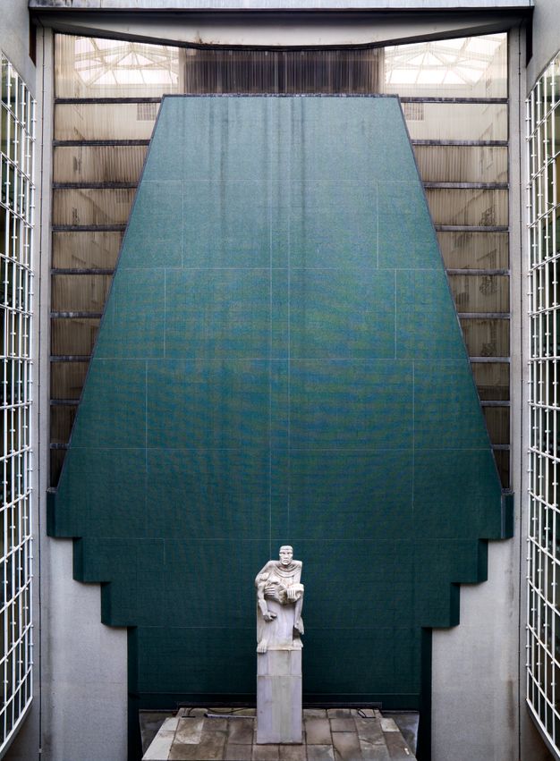 Architecture. An alcove is backed by blue stone that flares down from the top and then steps inwards to narrow at the bottom around a person holding the limp body of another.